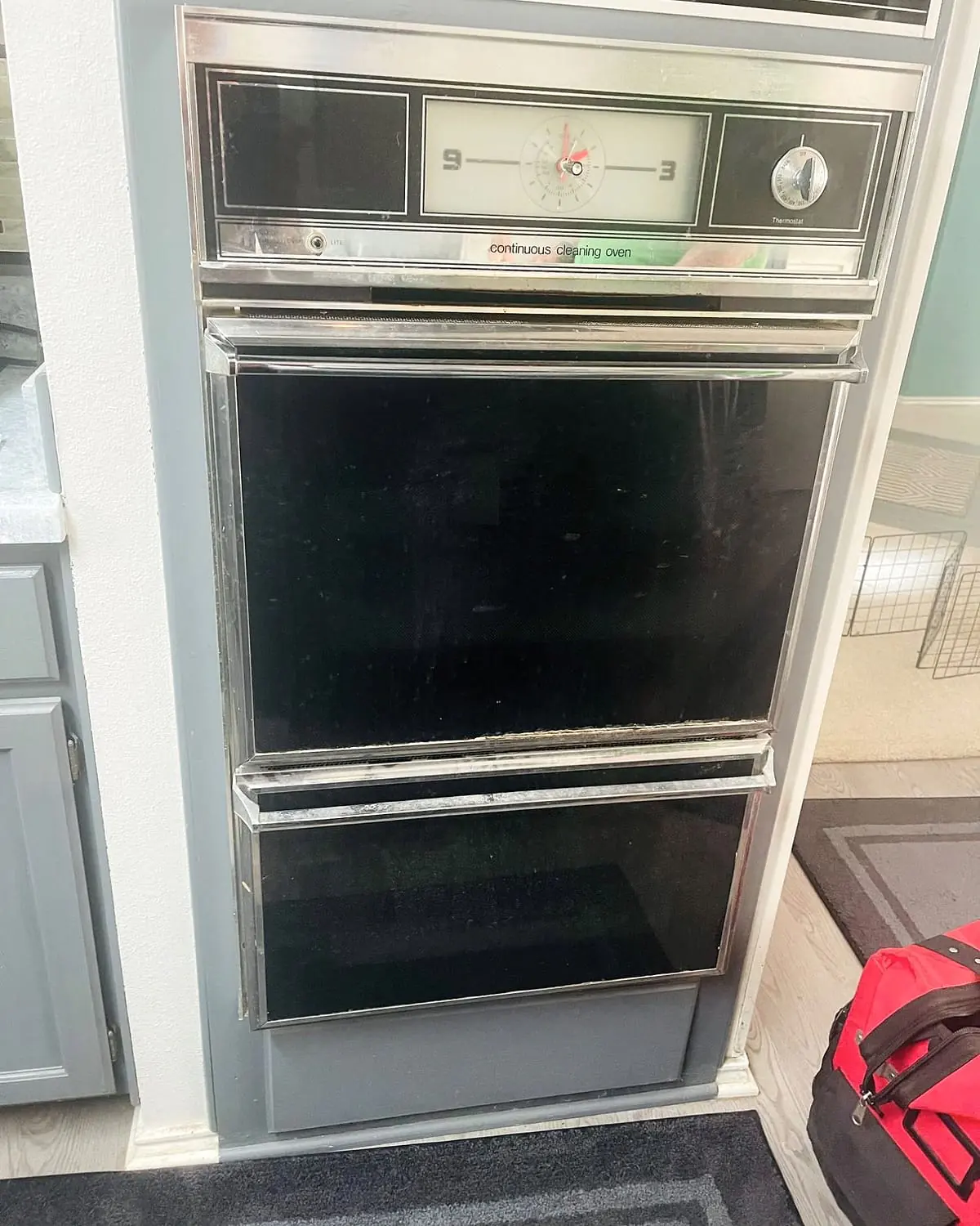 Older built-in double wall oven with a dial clock and thermostat, a red tool bag on the floor beside it